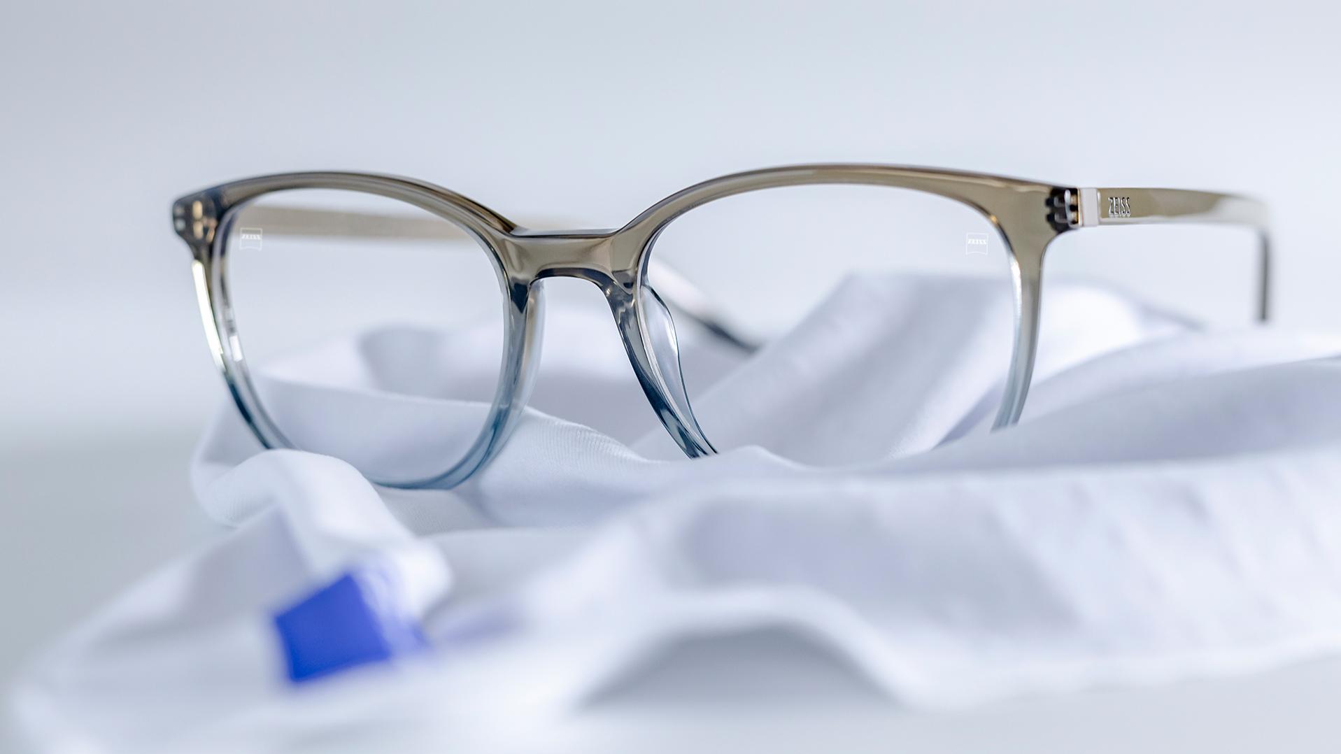 Pár okuliarov so sivomodrými rámami a šošovkami ZEISS s povrchom DuraVision® je položený na bielej handričke z mikrovlákna.