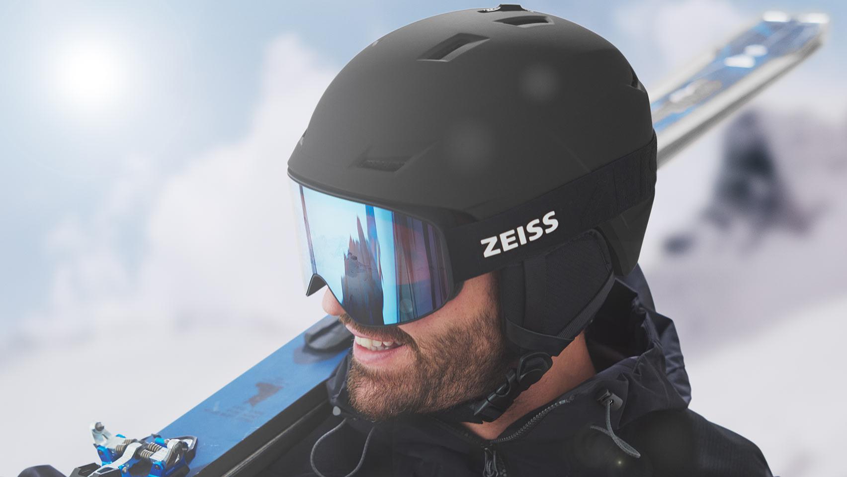 Muž má nasadenú čiernu prilbu s modrými zrkadlovými lyžiarskymi okuliarmi ZEISS, má lyže cez pravé rameno.
