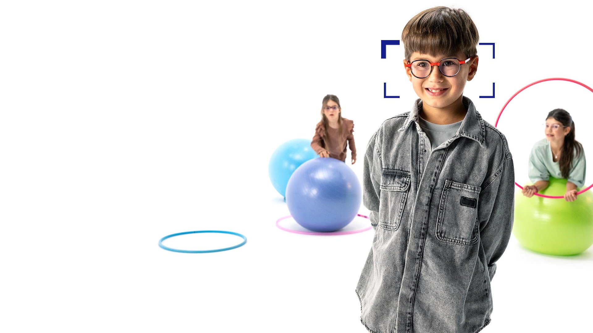 V popredí stojí hnedovlasý chlapec s okrúhlymi okuliarmi s šošovkami ZEISS MyoCare a smeje sa do fotoaparátu. V pozadí sú dve dievčatá so šošovkami ZEISS MyoCare hrajúce sa s obručami a gymnastickými loptami.
